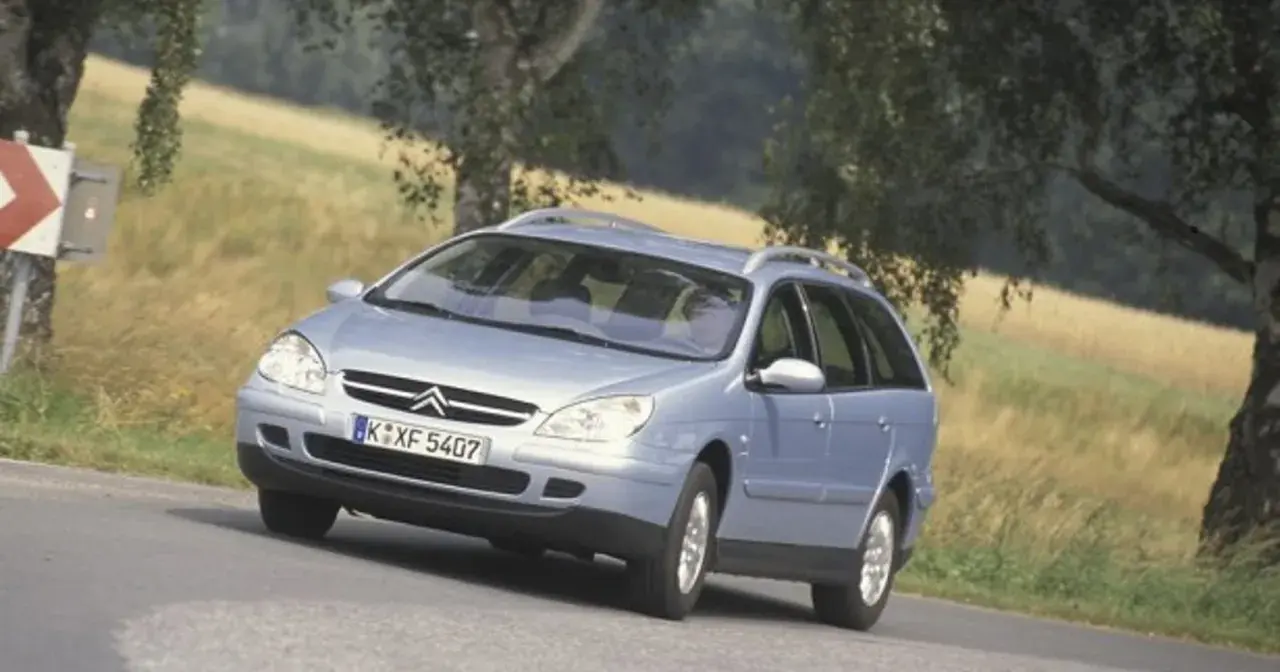 Jasnoniebieskie kombi Citroen na zakręcie drogi, z drzewami i polem w tle.