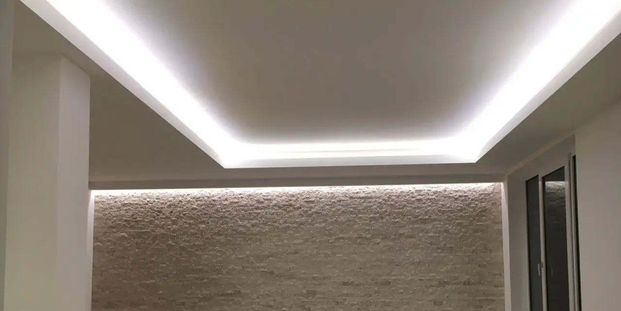 Illuminazione indiretta su velette in cartongesso, che creano un effetto soffuso su una parete in pietra chiara.