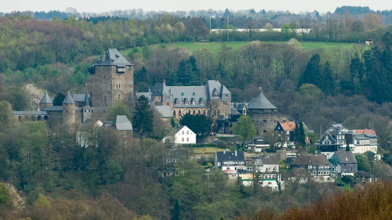 Zamek Schloss Burg w Solingen, otoczony zielenią, z wieżami i budynkami mieszkalnymi u podnóża.