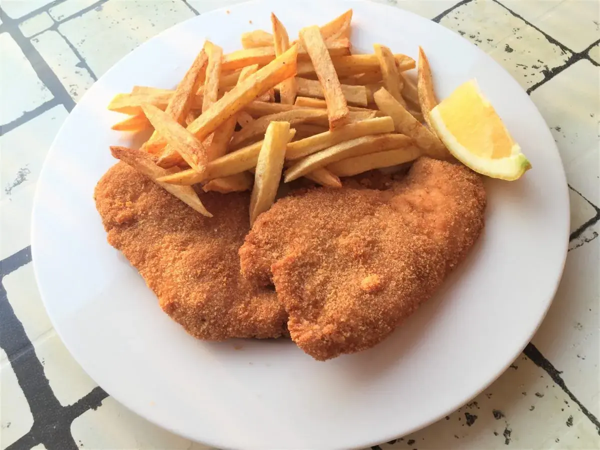 Due cotolette di pollo dorate e croccanti, accompagnate da patatine fritte e uno spicchio di limone su un piatto bianco.