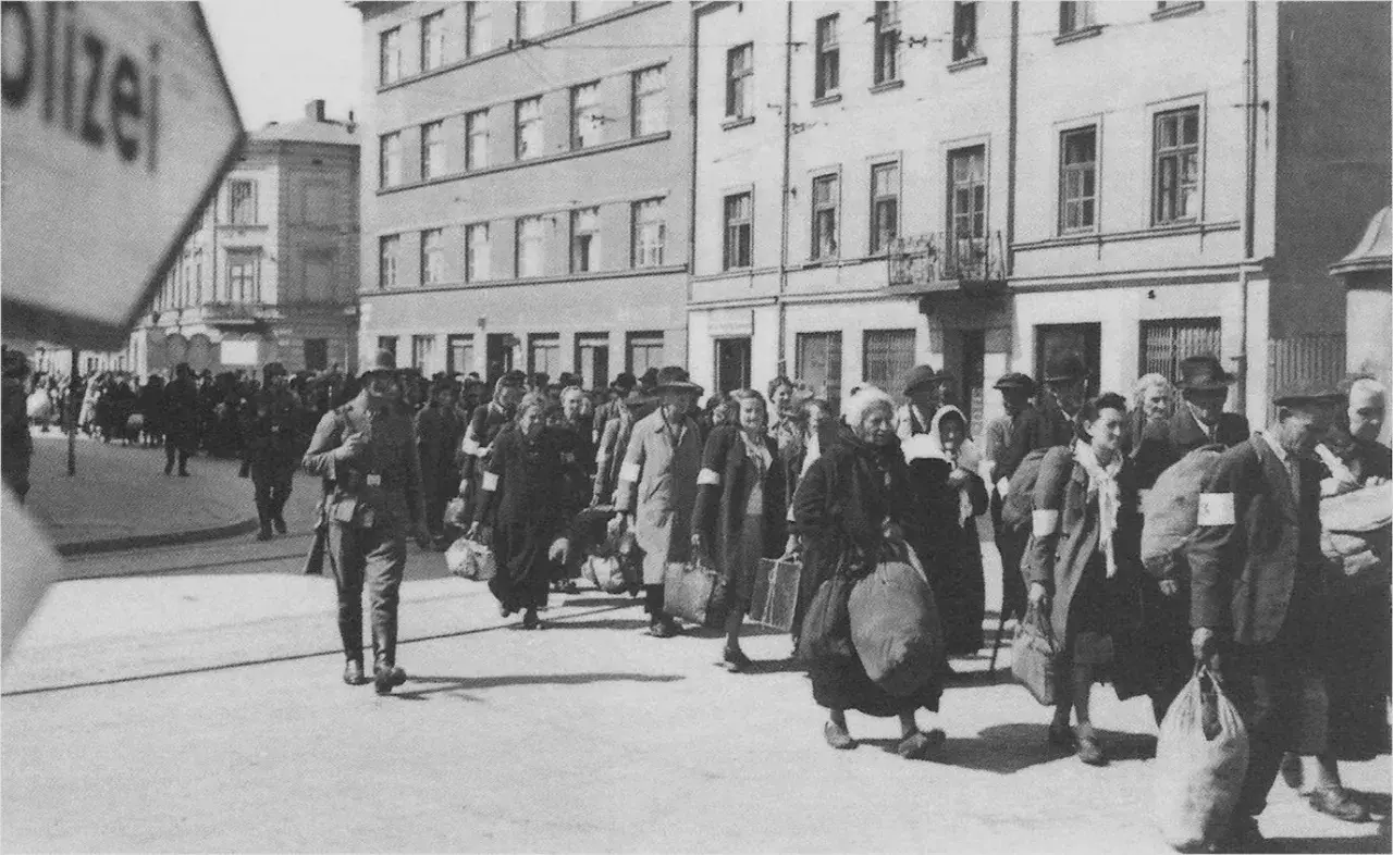 Människor med armbindlar ledsas förbi Schindlers fabrik. En soldat går framför.
