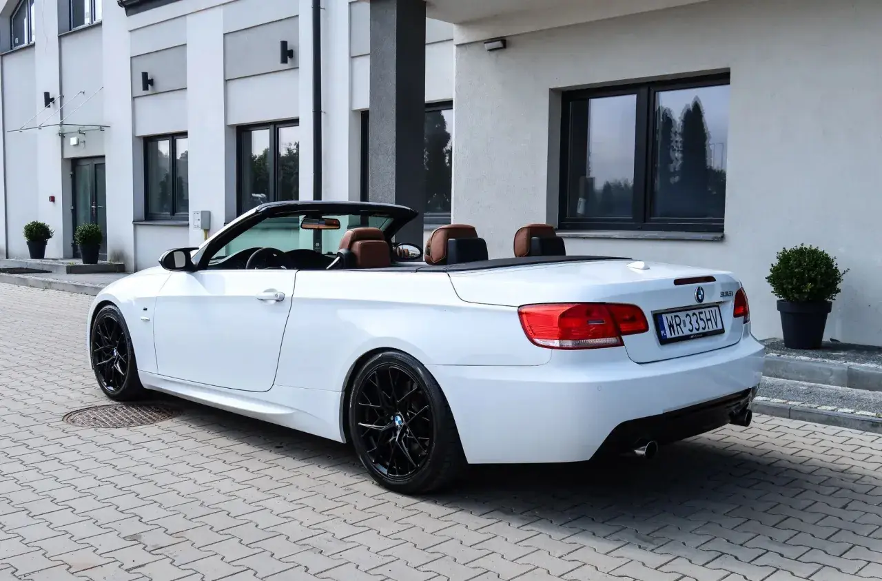 Białe BMW E93 cabrio z brązową skórzaną tapicerką i czarnymi felgami stoi na kostce brukowej przed nowoczesnym budynkiem.