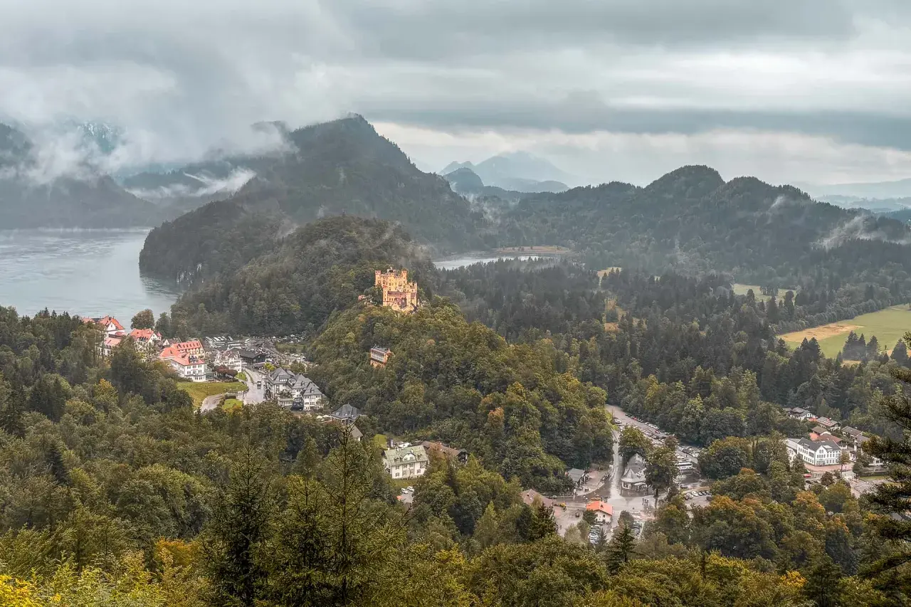 Schlechtes Wetter trübt den Urlaub. Ein gelbes Schloss thront auf einem bewaldeten Hügel über einem Dorf am See.