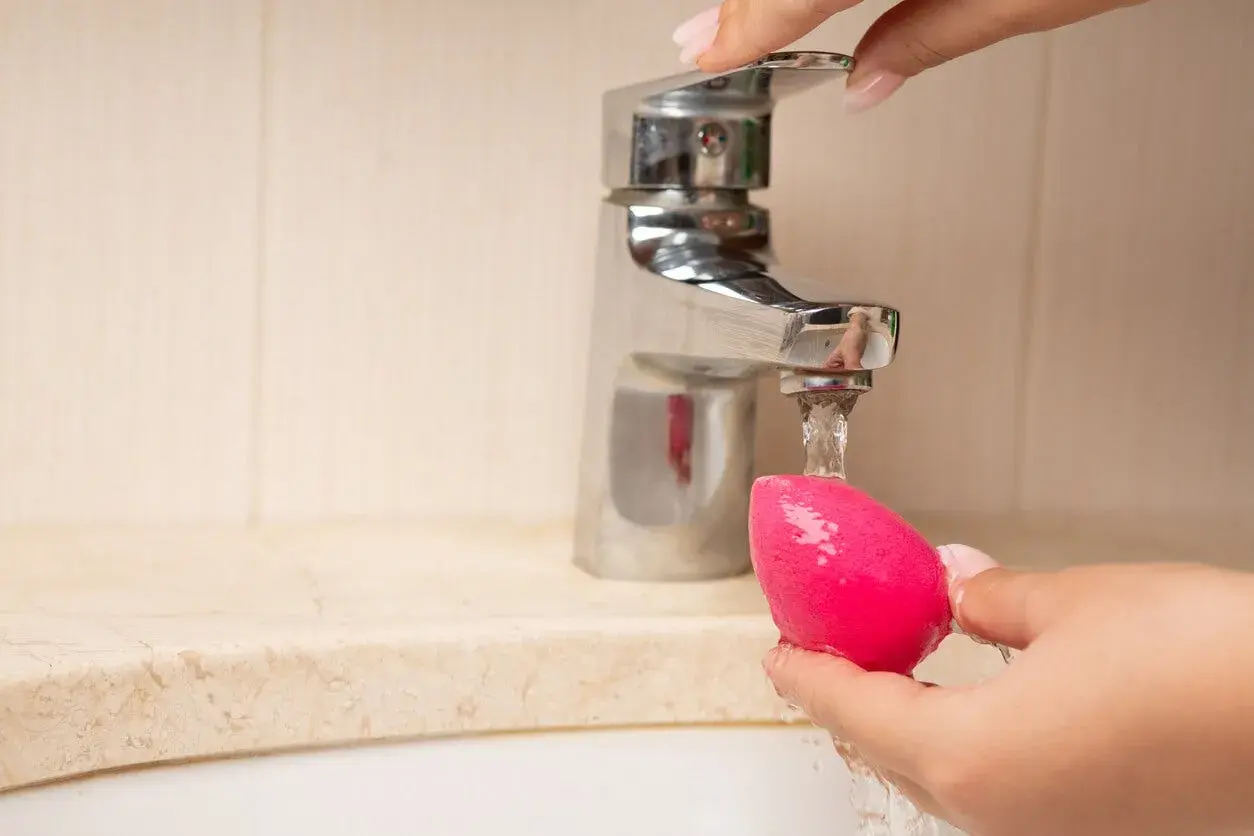 Mano limpiando esponja de maquillaje rosa bajo un grifo. Aprende como limpiar las esponjas de maquillaje para un mejor acabado.