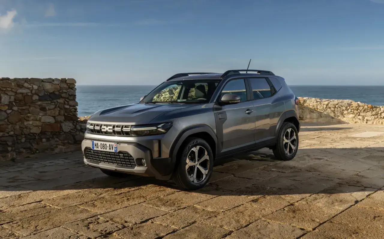Nowy Dacia SUV na tle kamiennego muru i morza.
