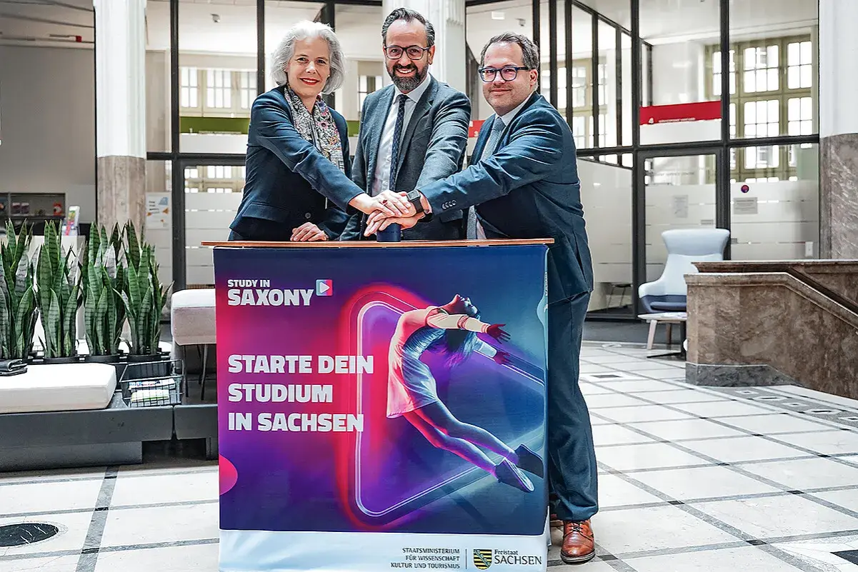 Drei Personen legen ihre Hände auf ein Banner für ein berufsbegleitendes Studium in Leipzig.