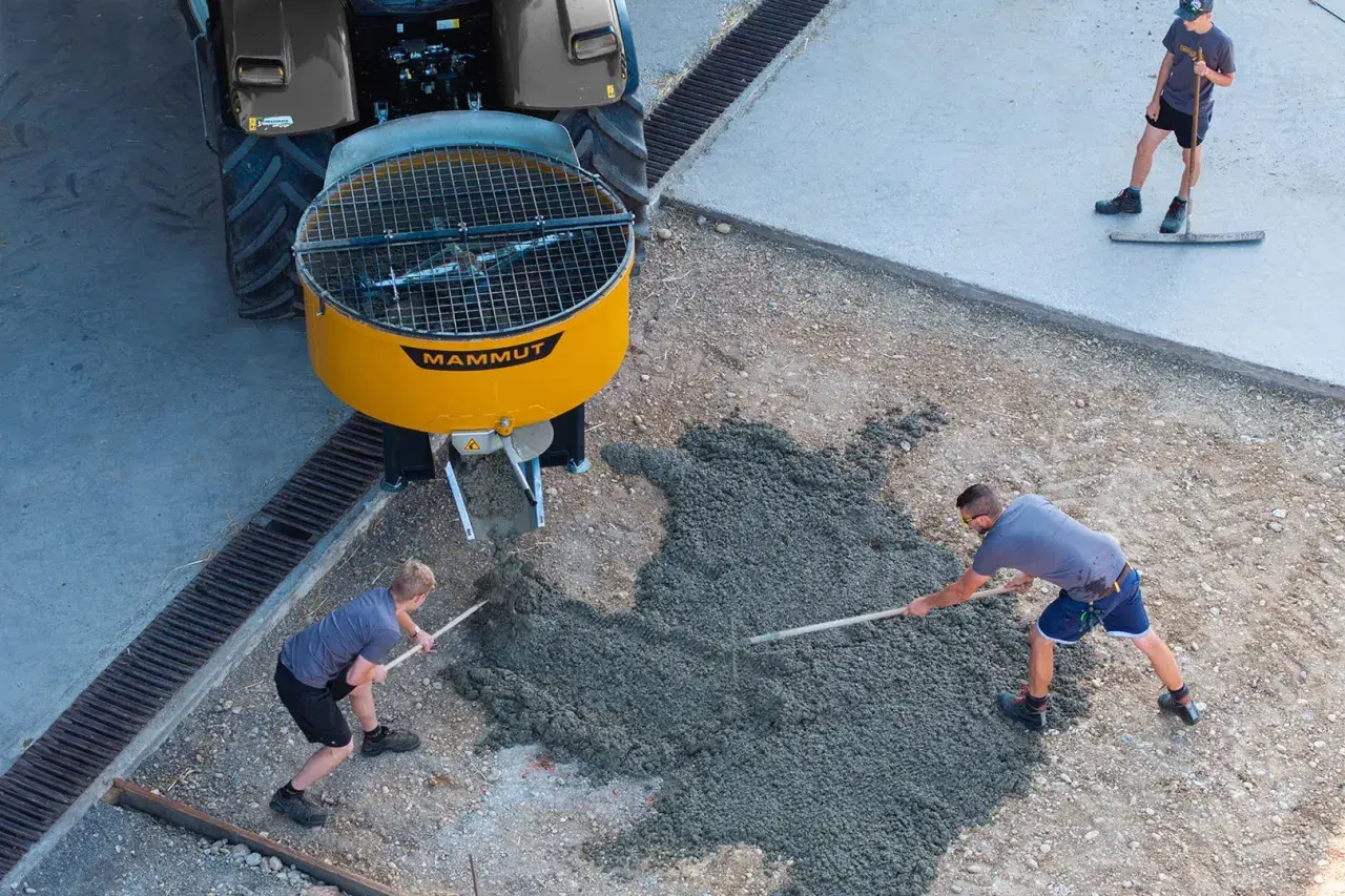 Zwei Arbeiter verteilen frischen Beton, der aus einem gelben Zwangsmischer einer Mammut-Maschine fließt. Ein dritter Arbeiter glättet den Beton.
