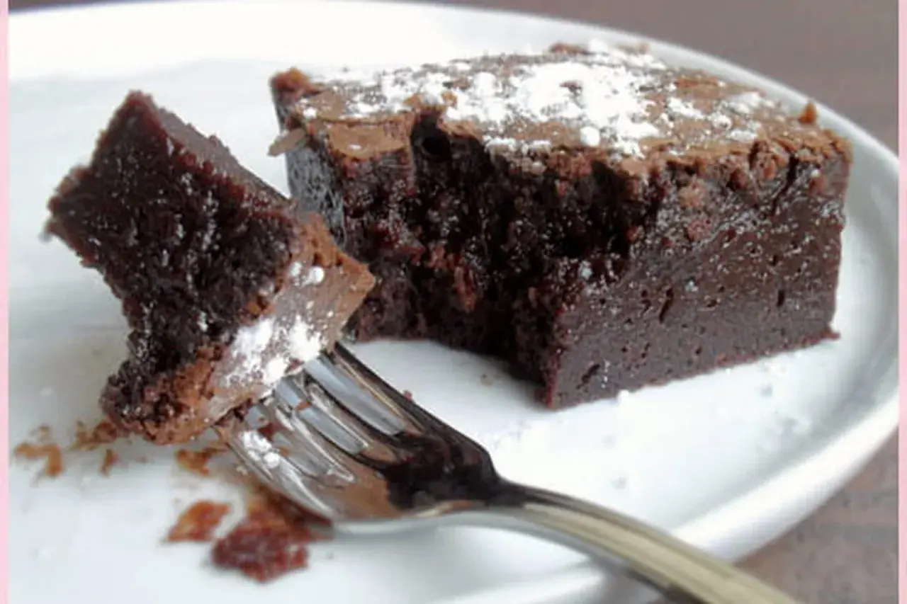 Un morceau de gâteau chocolat, saupoudré de sucre glace, est sur une assiette blanche. Une fourchette est prête à déguster ce délice.