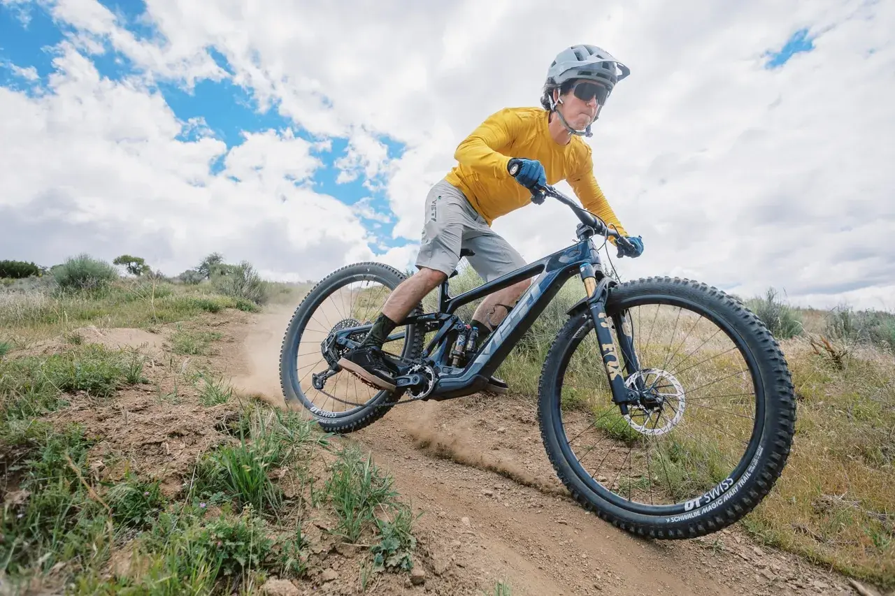 A mountain biker shreds a dusty trail on a Yeti MTE bike, kicking up dirt under a cloudy sky.