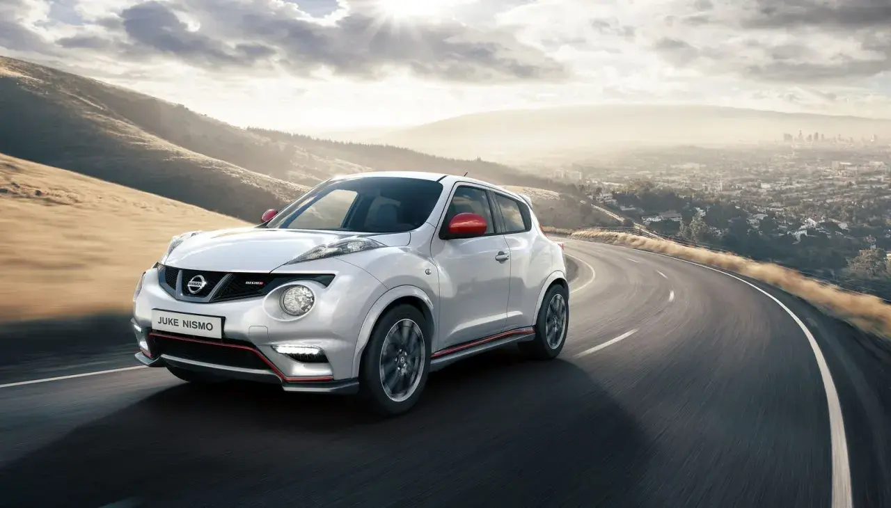 Biały Nissan Juke Nismo z czerwonymi lusterkami pędzi po krętej drodze z widokiem na miasto w oddali.