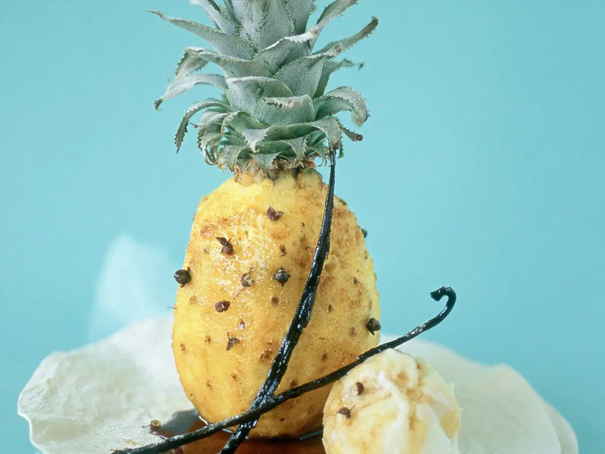 Un ananas rôti, parsemé de clous de girofle et de gousses de vanille, sur un fond bleu pâle. Un délice sucré et épicé.