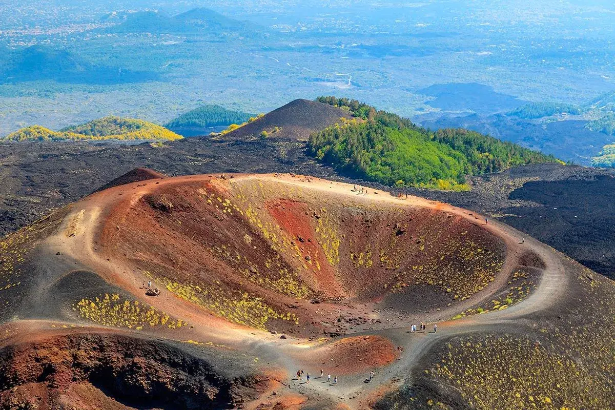 Widok na krater wulkanu Etna, gdzie ścieżka prowadzi turystów na wejście na etnę. W oddali zielone wzgórza i miasto.