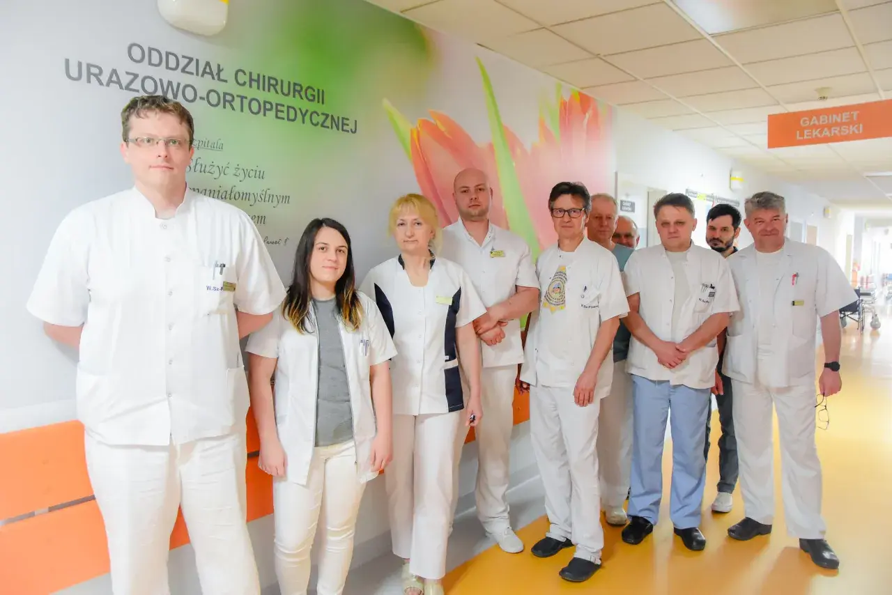 Grupa lekarzy i pielęgniarek w białych fartuchach stoi w szpitalnym korytarzu.