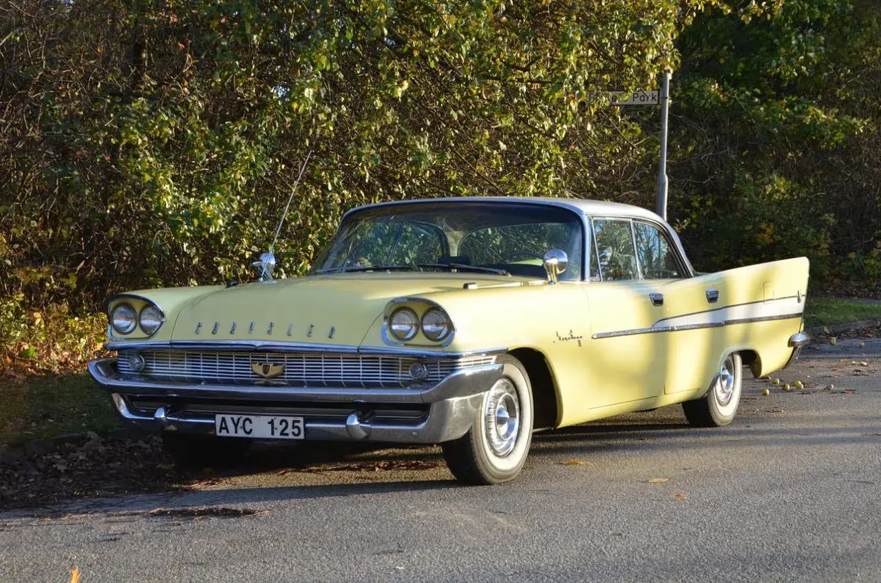 Gul Chrysler Saratoga från 1950-talet, med kromdetaljer och vita däcksidor, parkerad vid en lummig buske.
