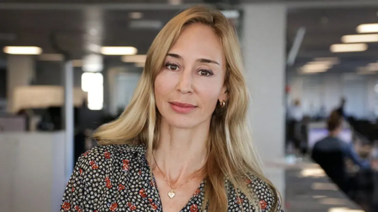 A blonde woman with a floral shirt and a heart necklace smiles at the camera in an office setting.