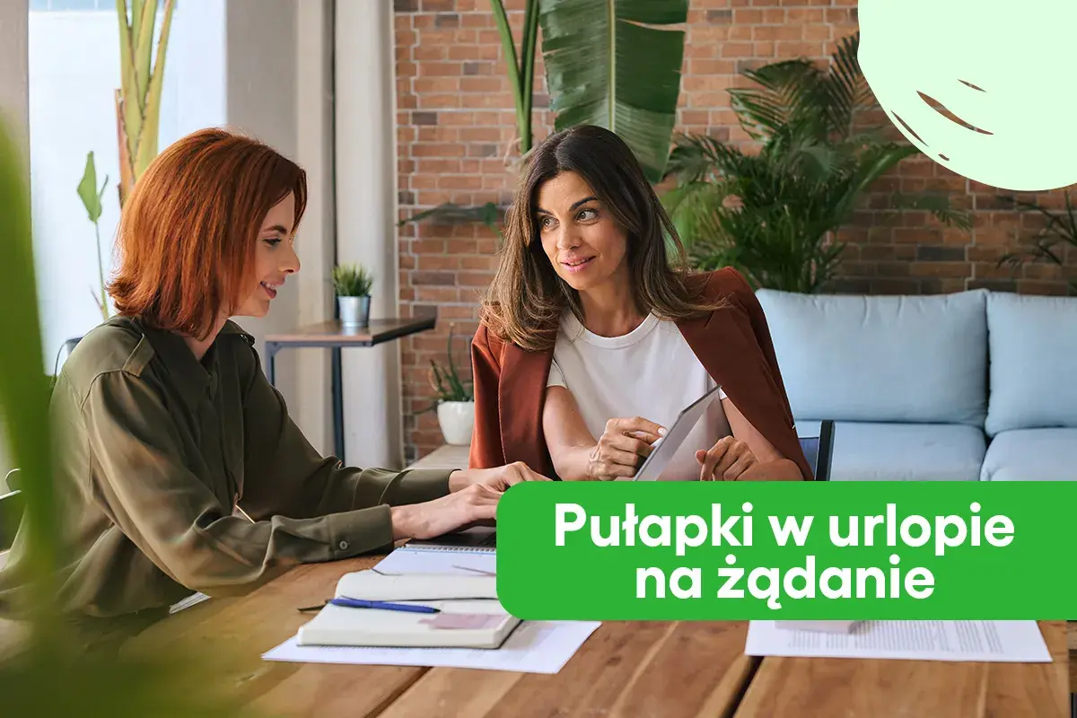 Dwie kobiety rozmawiają przy biurku, jedna pokazuje coś na tablecie.