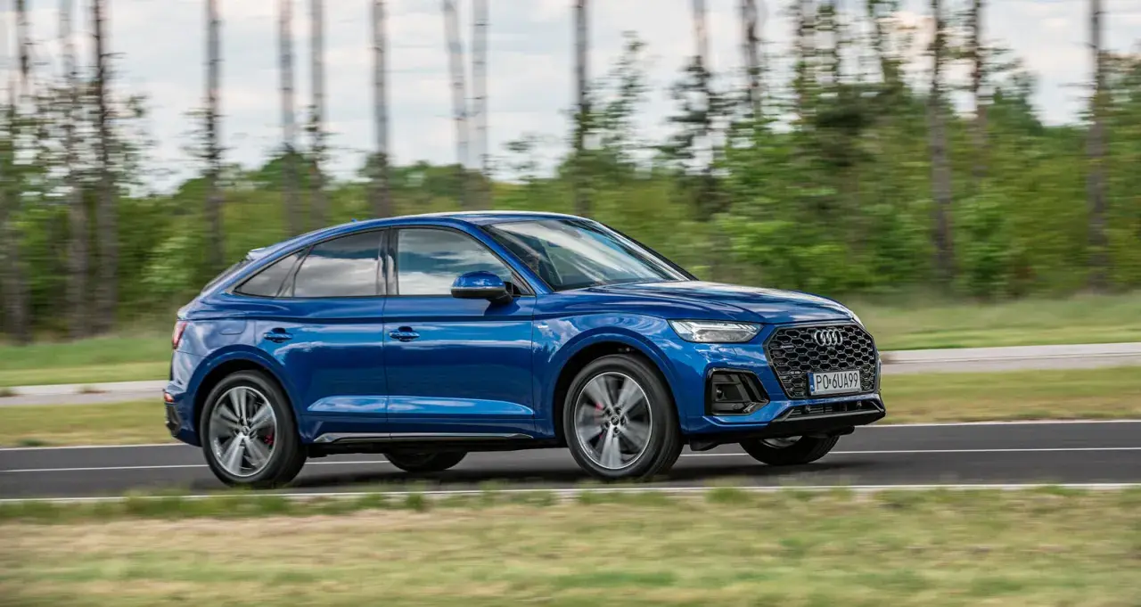 Błękitny Audi Q5 Sportback 2025 pędzi po drodze, otoczony zielenią i drzewami.