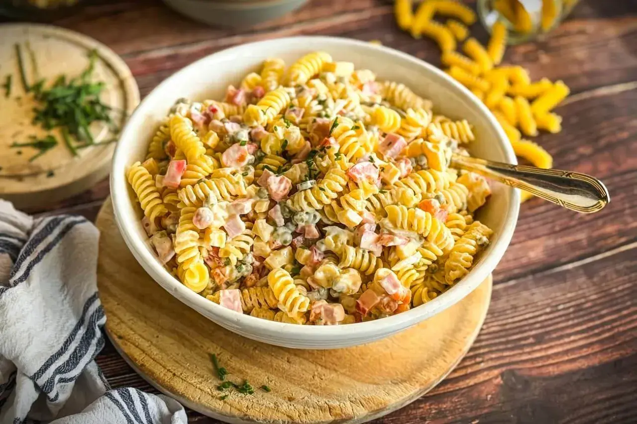 Schüssel mit cremigem Nudelsalat, gefüllt mit Schinken, Erbsen und Kräutern. Ein Löffel liegt darin. Dieser ist der beste Nudelsalat!