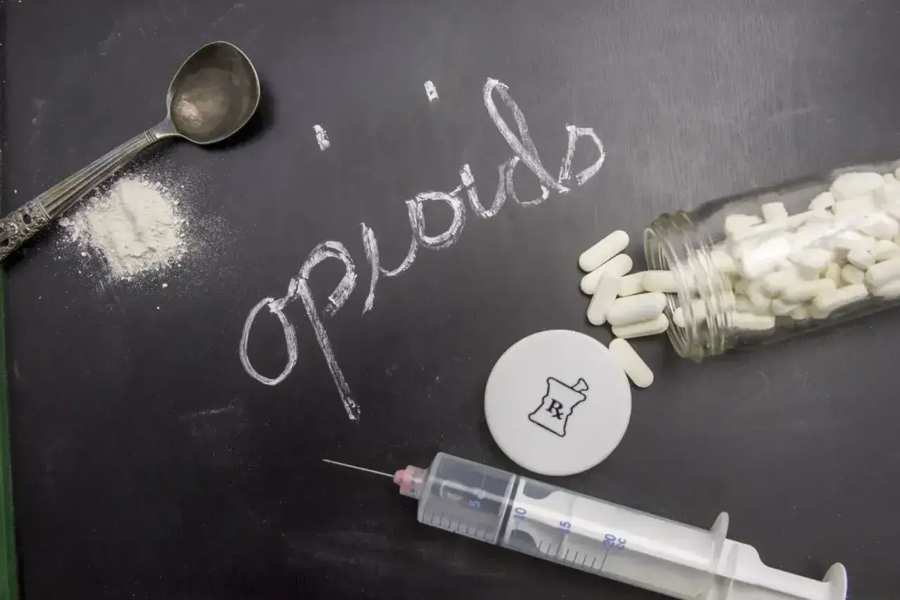 Na czarnej tablicy napis "opioids" kredą. Obok łyżka z proszkiem, strzykawka, fiolka z kapsułkami i symbol apteczny.