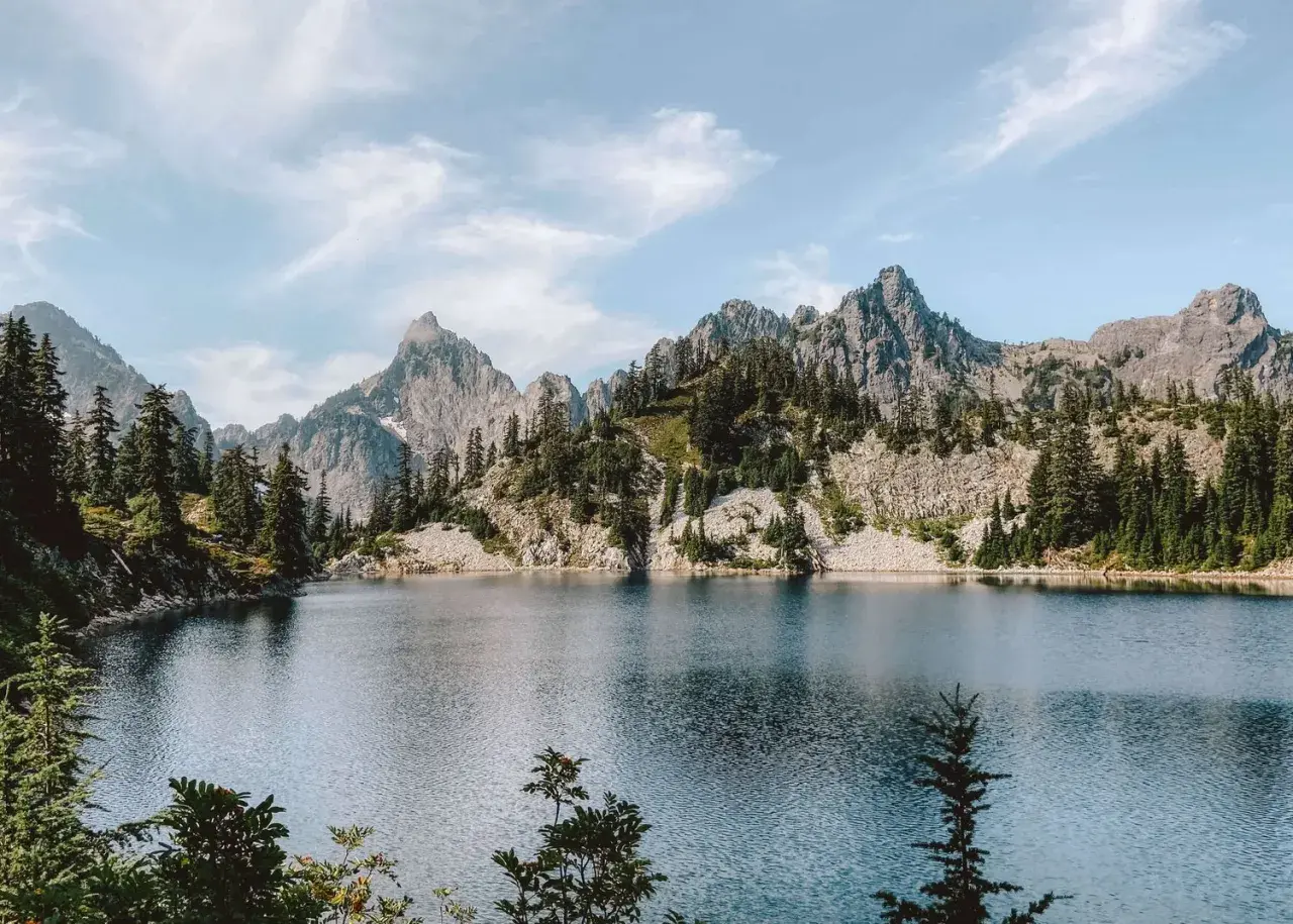 A serene alpine lake reflects jagged, tree-covered mountains. This view makes you wonder how long to hike 16 miles to reach such a peaceful spot.