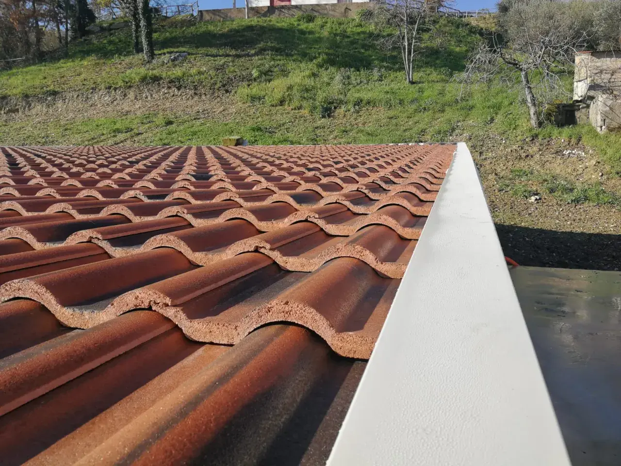 Tegole per tetti a bassa pendenza, color terracotta, disposte in file ordinate su un tetto. Sullo sfondo, un pendio erboso.