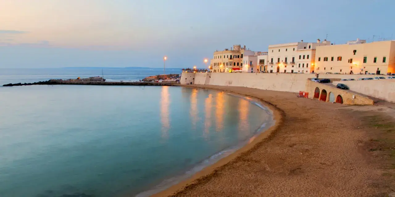 La costa di un paese vicino a Lecce al tramonto, con edifici bianchi, una spiaggia sabbiosa e il mare calmo.