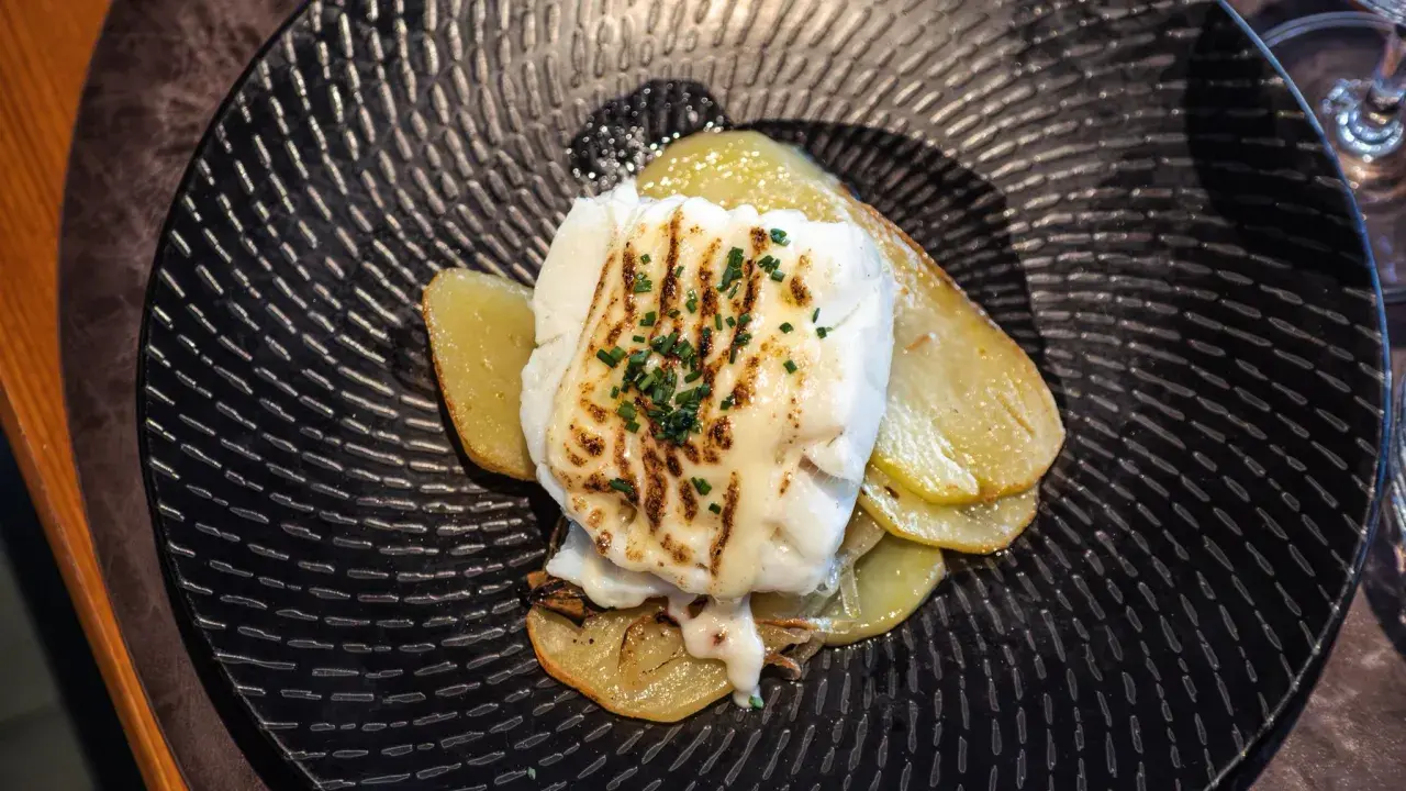 Bacalao gratinado con alioli, servido sobre patatas finas en un plato negro texturizado.