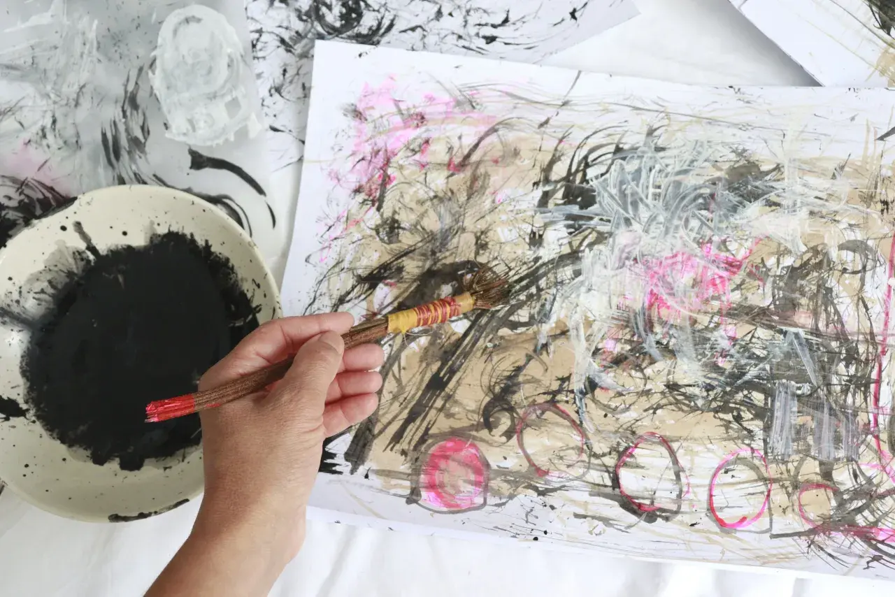 A hand holds a brush, adding bold strokes to an abstract drawing. Black ink splatters a palette beside the energetic artwork.