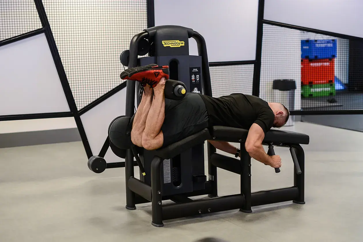 Mężczyzna wykonuje ćwiczenie na maszynie do uginania nóg leżąc (hamstring curl), budując siłę tylnej części uda.