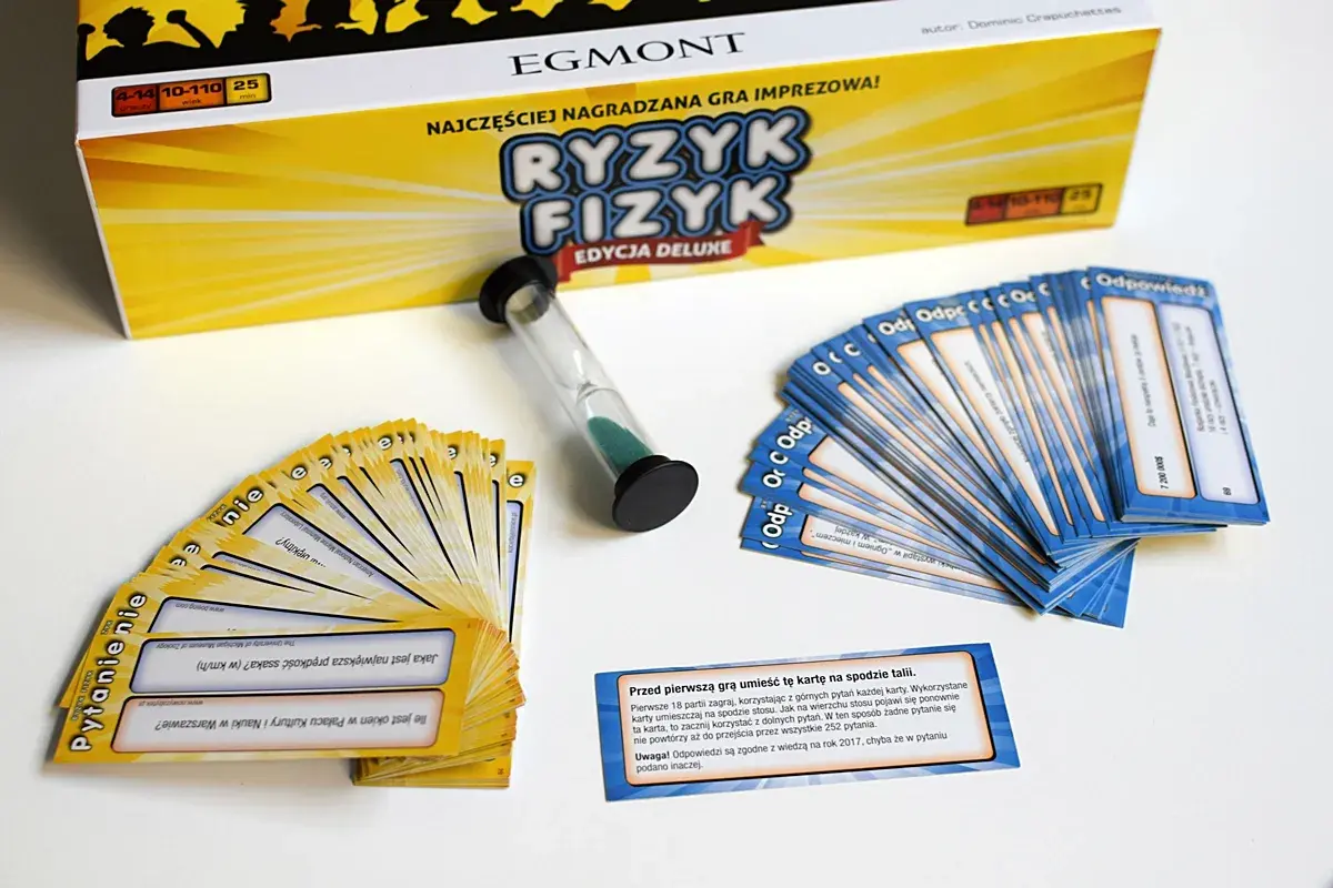 Pudełko gry "Ryzyk Fizyk" i rozłożone karty pytań i odpowiedzi. Czasomierz piaskowy czeka na rozpoczęcie rundy.