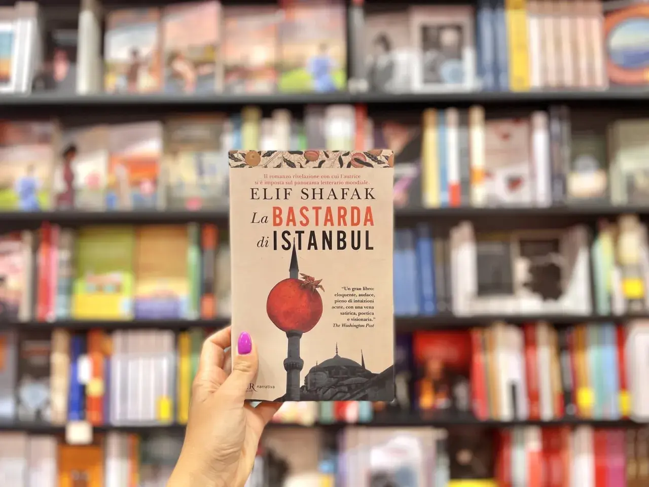 Mano tiene "La Bastarda di Istanbul", un libro che esplora le storie di Istanbul, tra scaffali pieni di libri.