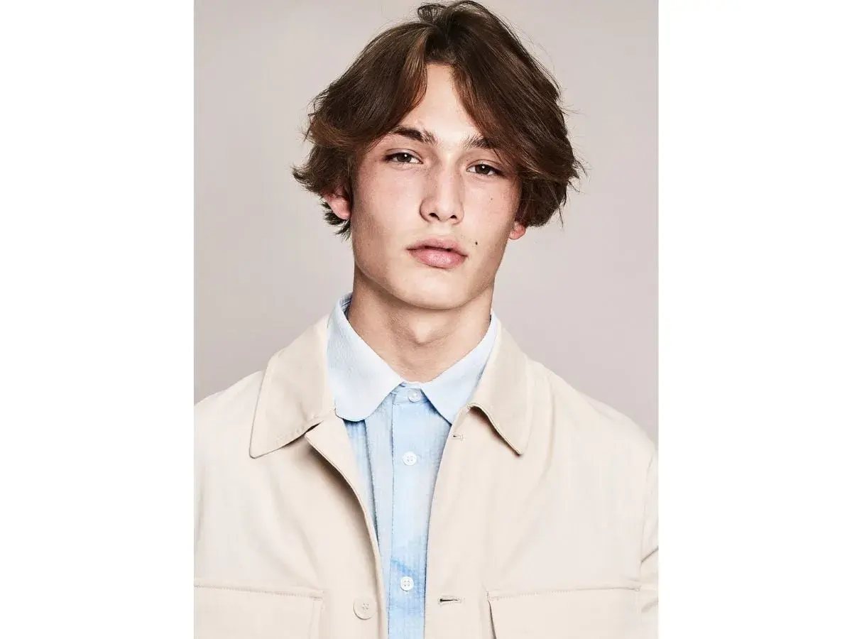 Ragazzo con capelli castani mossi e acconciature alla moda, indossa una camicia azzurra e una giacca beige.