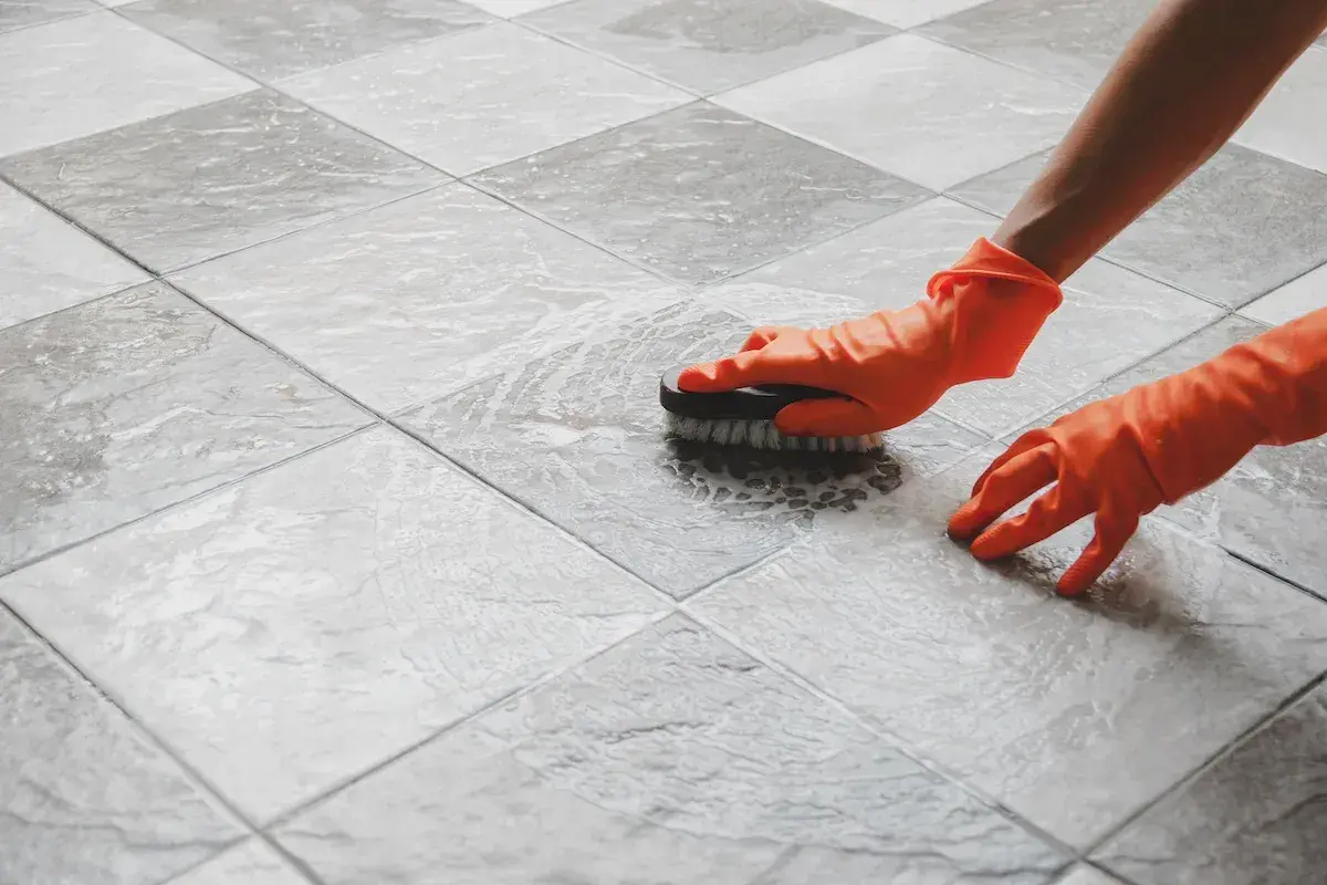 Mani con guanti arancioni puliscono un pavimento piastrellato con una spazzola, mostrando come pulire il pavimento dopo la posa.