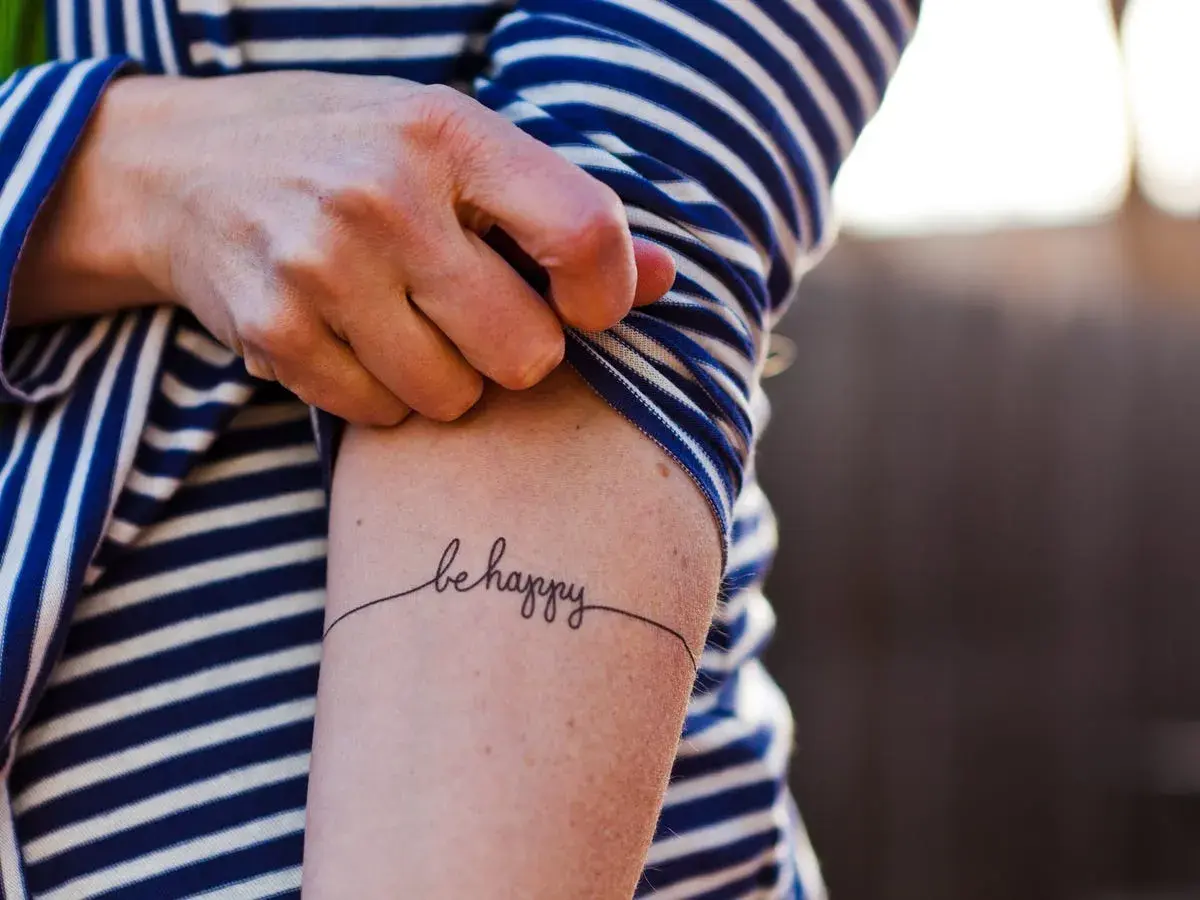 Un brazo con una camiseta a rayas azules y blancas muestra un delicado tatuaje de caligrafía que dice "be happy", una forma de arte para transformar cicatrices.