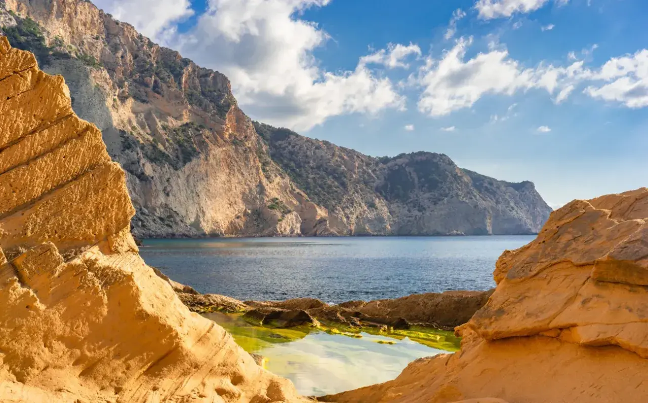 Rocas ocres en primer plano en sa Pedrera Ibiza, con el mar azul y montañas escarpadas al fondo bajo un cielo nublado.