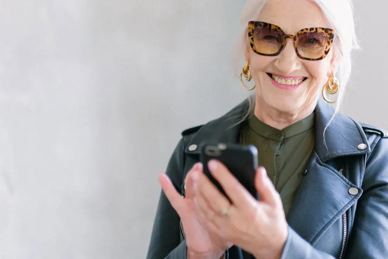 Uśmiechnięta starsza pani w okularach w panterkę sprawdza telefon. Może szuka informacji o kodeksie pracy i odprawie emerytalnej.