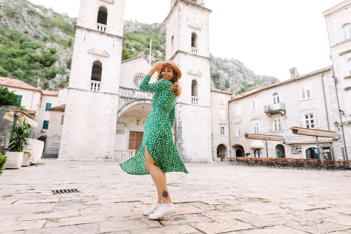 Une femme porte une robe verte fleurie et des baskets blanches, un chapeau de paille sur la tête, dans une place pavée devant une église.