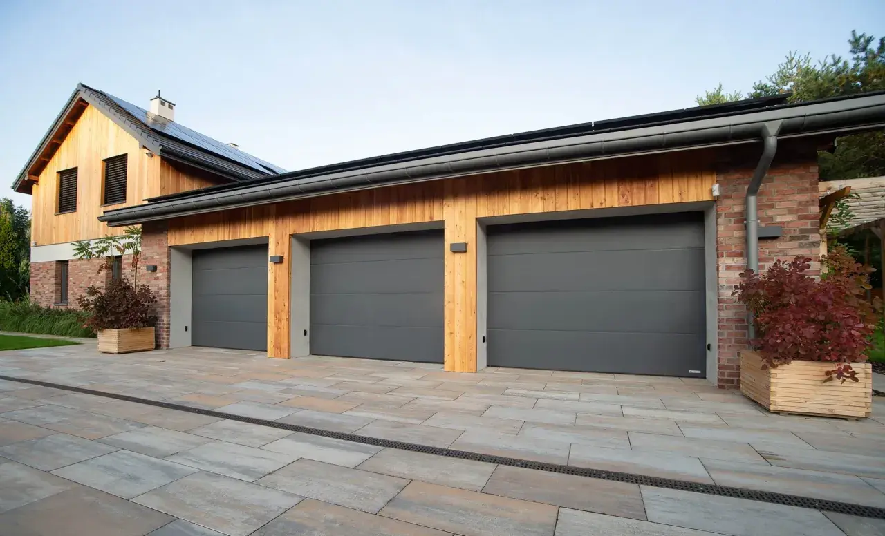 Tre moderna garageportar i mörkgrått framför ett hus med träfasad och tegelfasad.