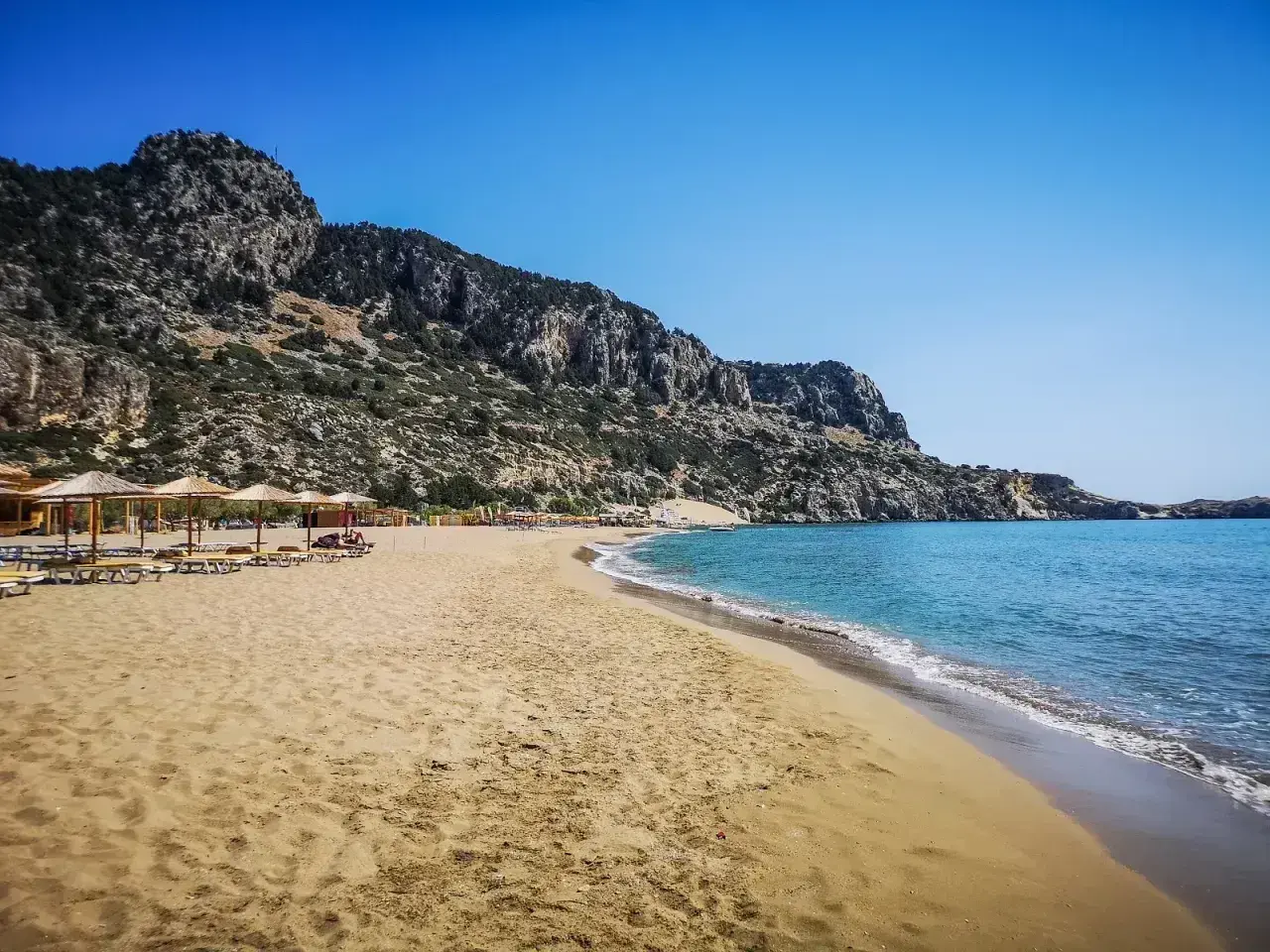 Piękna, piaszczysta plaża Tsambika z turkusową wodą i skalistym wzgórzem w tle. Leżaki i parasole czekają na plażowiczów.