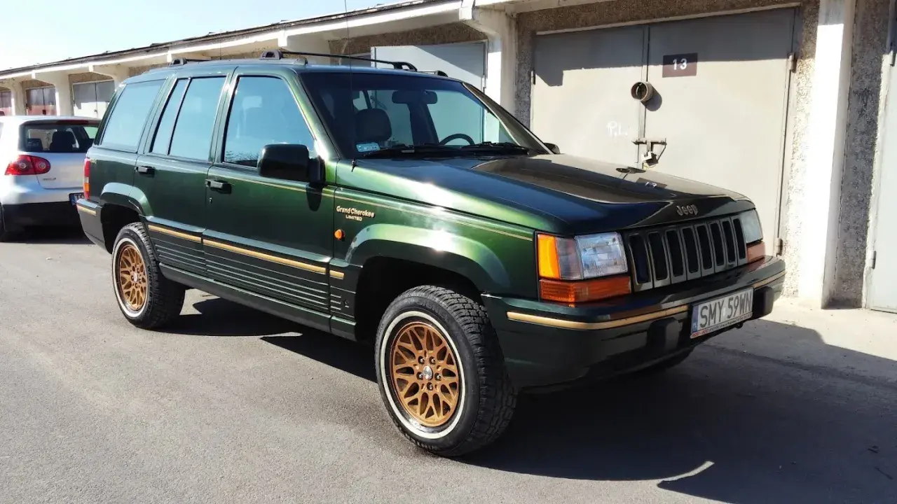 Zielony Jeep Grand Cherokee Limited z lat 90. na złotych felgach, zaparkowany przed garażami.