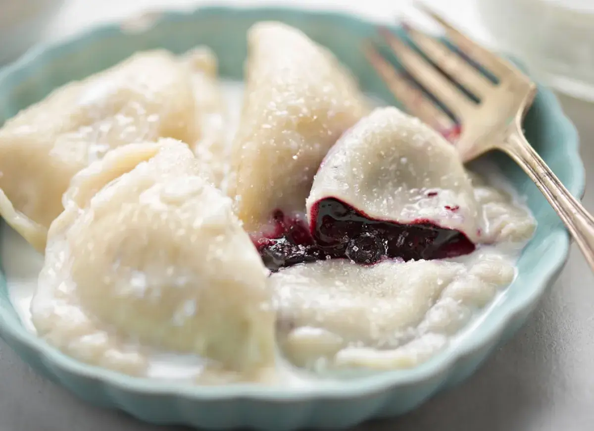 Pyszne pierogi z jagodami, polane śmietaną i posypane cukrem, czekają na widelca.