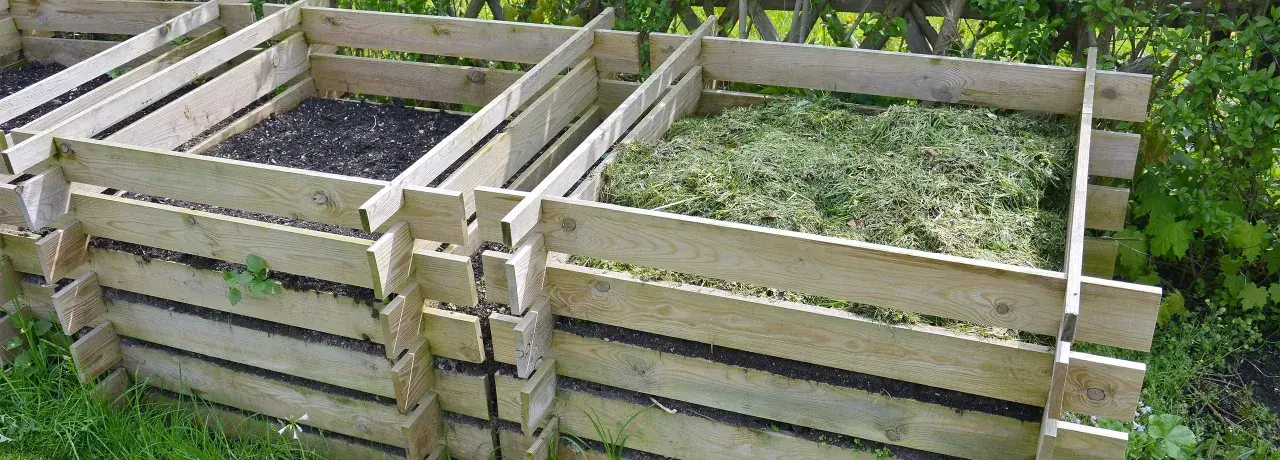 Holzkomposter mit zwei Fächern, eines gefüllt mit Erde, das andere mit frisch gemähtem Gras, bereit zum Kompost zerkleinern.