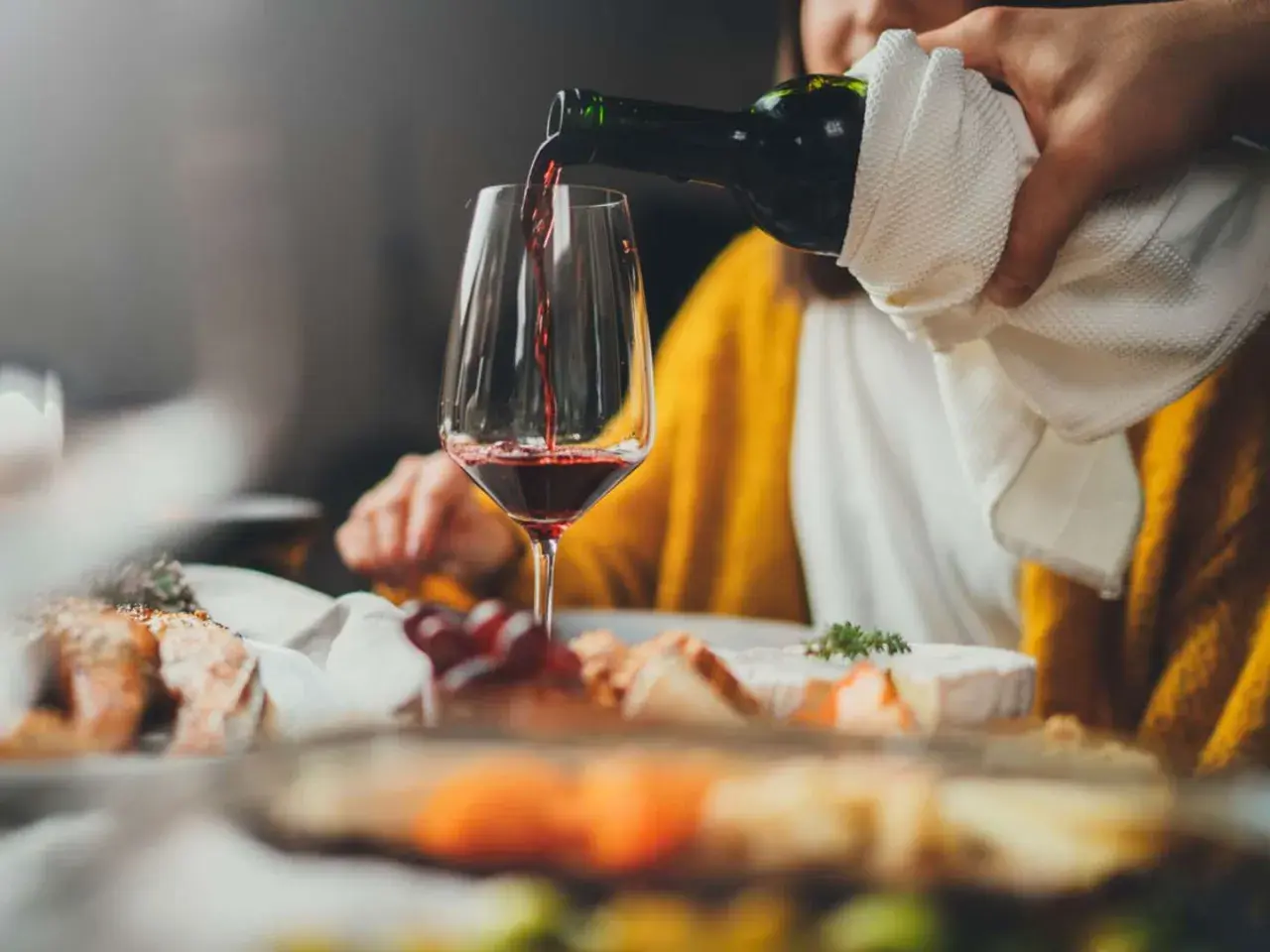 Rödvinet hälls upp i ett glas vid en middagsbjudning. En person i gul tröja sitter bakom. Frågan om hur mycket vin per person är relevant vid sådana tillfällen.