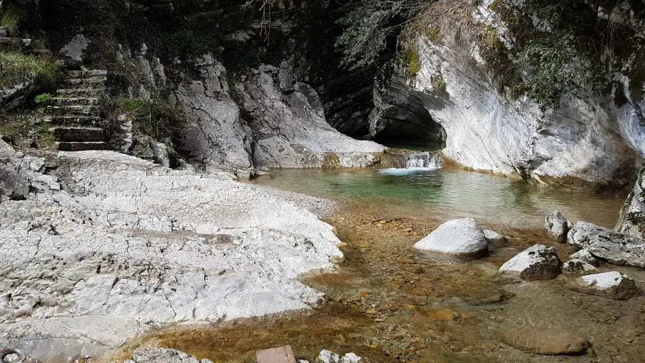 Sentiero di pietra conduce a un ruscello cristallino con piccole cascate di piovere, incastonato tra rocce bianche e vegetazione.