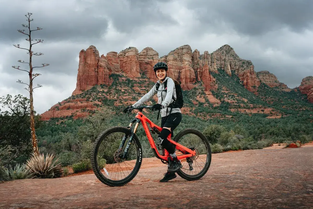 A smiling woman in riding gear poses on her bright orange mountain bike, ready for a trail tune-up amidst the stunning red rock formations of Sedona.