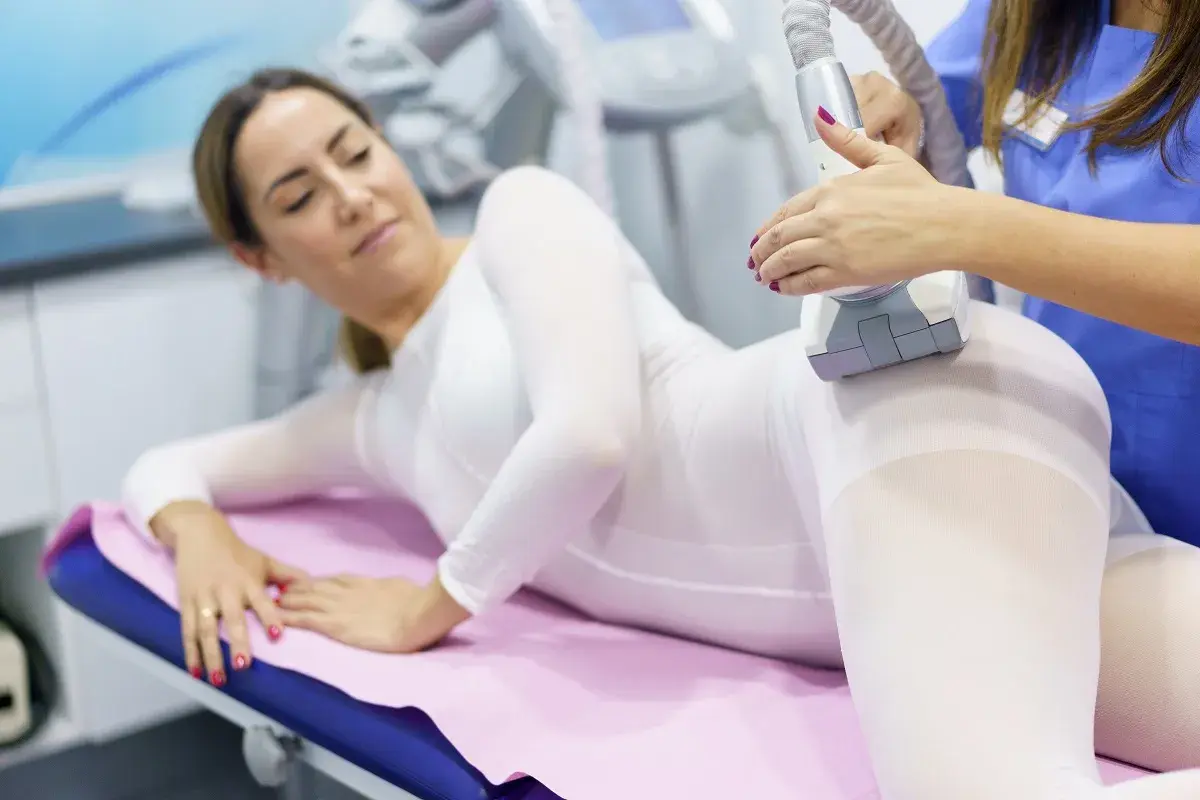 Kobieta w białym kombinezonie leży na kozetce, a kosmetyczka wykonuje zabieg endermologii. To nowoczesny icoon piękna.