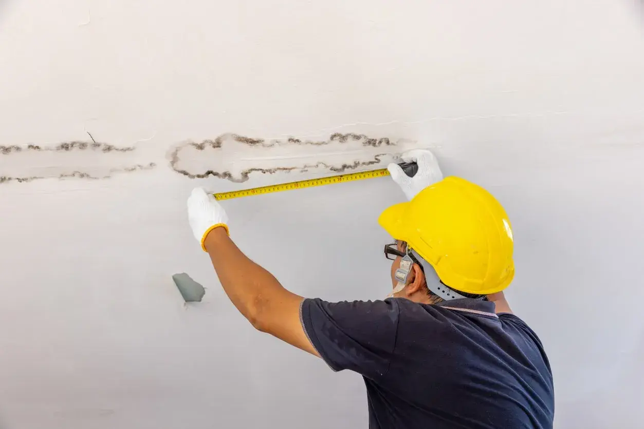 Un homme en casque jaune mesure des taches d'humidité au plafond, cherchant une solution humidité maison.