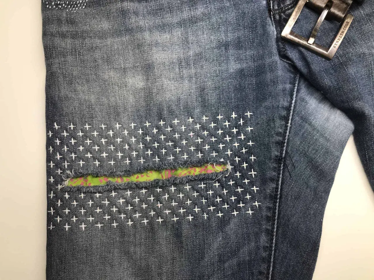 Close-up of denim jeans with a colorful fabric patch and white cross-stitch embroidery, showcasing a stylish sashiko jeans repair.