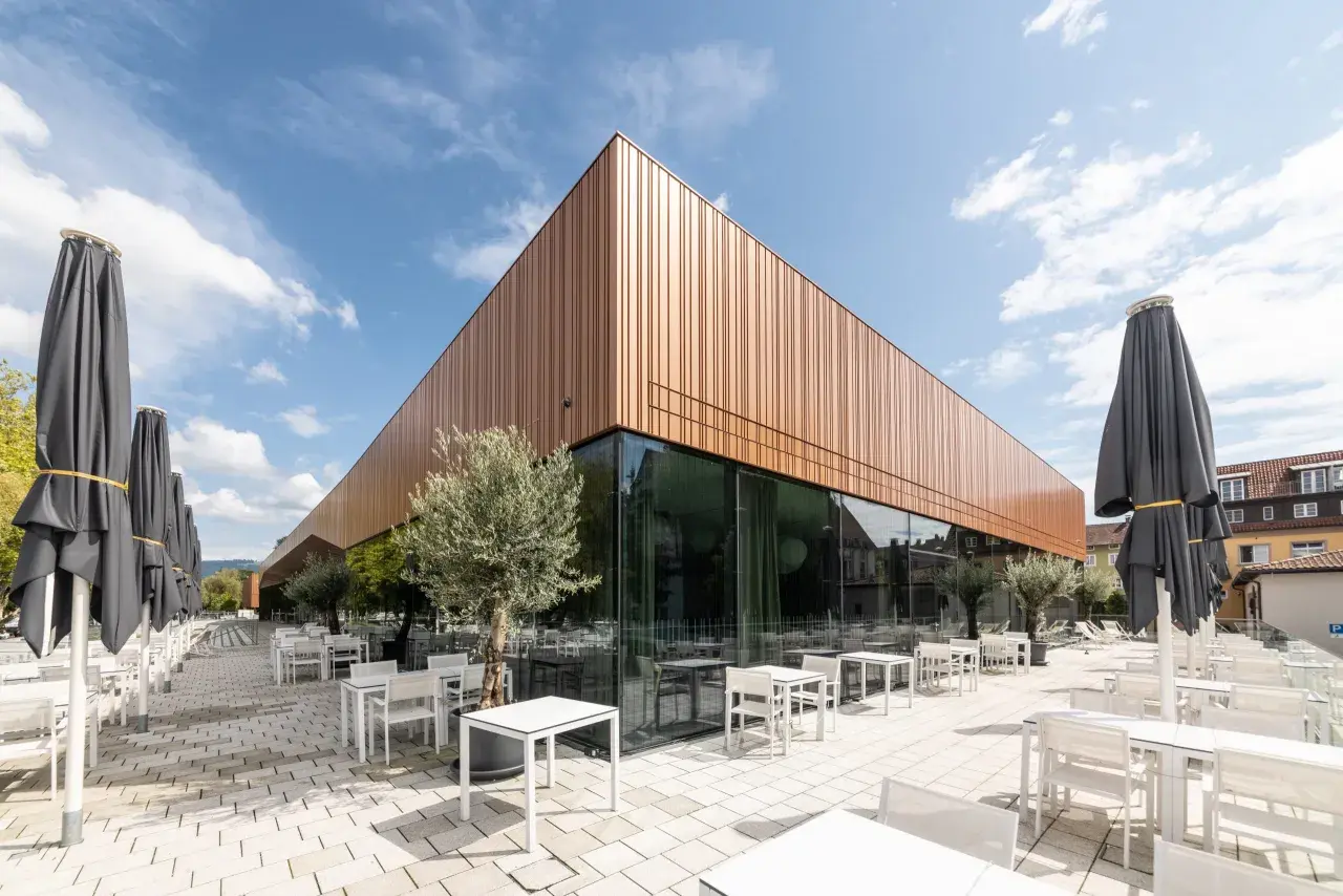Moderne Architektur der Inselhalle Lindau mit kupferfarbener Fassade, Glasfront und vielen leeren Tischen und Stühlen auf einer gepflasterten Terrasse.