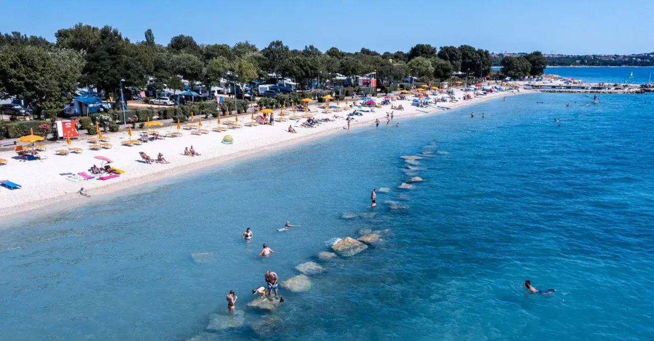 Słoneczna plaża z turystami, parasolami i kamperami w tle. Woda kusi do kąpieli, a kamienie tworzą naturalne falochrony. Idealne miejsce na bi village camping.